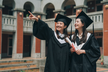 asian university college graduate student woman in graduation academic dress gown smiling pointing sky showing best friend with traditional red brick building. education or success hope concept.の写真素材