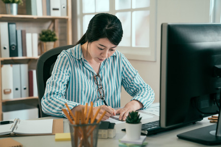 asian woman architect engineer design working planning concept. young chinese female sitting at desk in company against window indoors. elegant interior worker using ruler and pen drawing at computerの写真素材