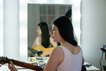 back view beautiful korean woman singer playing acoustic guitar sitting at vanity table in dressing room with applied makeup. young asian girl musician ready for show concert practicing in backstage.の写真素材
