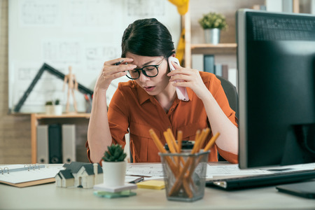 asian female engineer receiving bad news on cell phone. worried young girl architect care about her new construction eco friendly project hand on forehead think solution. woman frowning solve problemの写真素材