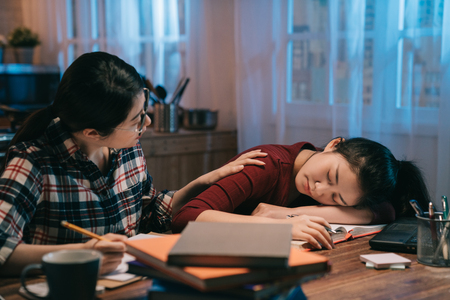 Two japanese girls students help each other doing homework together in midnight. one lady fell asleep another shouts at her trying to wake hands touching shoulder. hardworking education concept.の写真素材