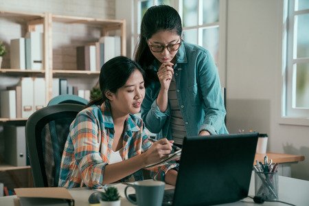 Young creative business people at office. two asian woman colleague in bright vintage studio talking on festival event plan on notebook and laptop pc against window. female coworker teach internの写真素材