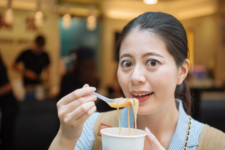 asian female travel backpacker looking camera eating vermicelli noodles famous shop located in taipei taiwan. young girl japanese tourist trying taiwanese tasty local specialty food on street vendorの写真素材