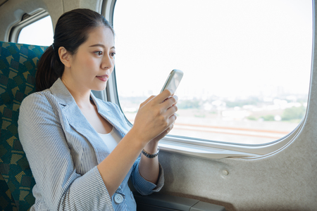 Businesswoman commuting to work on train using mobile phone sitting on seat by window. asian elegant office lady employee manager in suit texting message on cellphone connect internet while commuteの写真素材