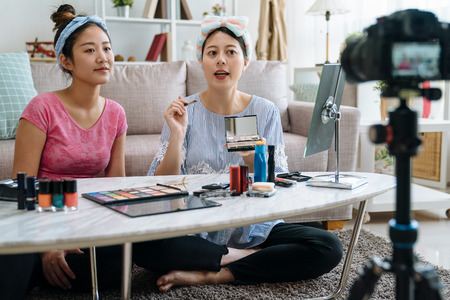 Beautiful asian woman blogger showing how to make up and use cosmetics. young girl vlogger friend together recording makeup tutorial video by professional camera in living room at home sit on carpetの写真素材