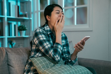 Sad asian girl in shirt yawning browsing boring websites on smartphone during leisure time in night apartment.の写真素材