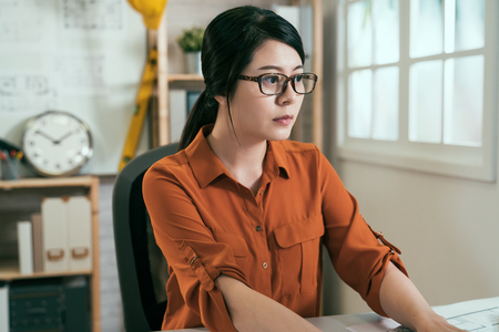serious concentrated asian female architect working in modern office. girl employee engineer sitting desk and doing creative new project of alternative energy building. woman in glasses use computerの写真素材