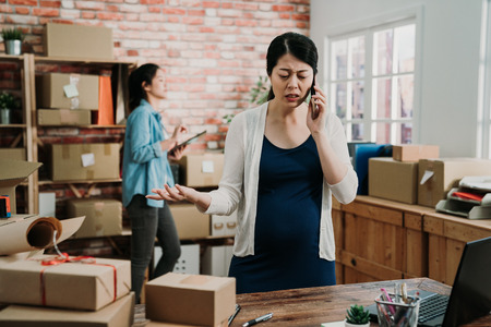 two startup small business female owners working at workplace. stressed freelance pregnant woman seller check product order for delivery to customer talking on smart phone argue angry with producer.の写真素材