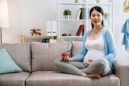 Pregnant asian young lady eating fresh fruits living healthy lifestyle taking care of child. elegant woman holding apple face camera smiling hands touch baby in belly sitting on couch sofa at home.の写真素材
