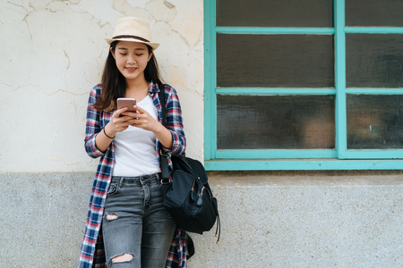 portrait of happy asian girl dressed in hat and jeans standing using mobile phone on city street against wall by old house window in town. smiling lady carry backpack texting on cellphone online smsの写真素材