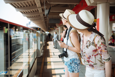 two young women friends travelers with backpack and camera  standing on outdoor platform in metro station looking for train.の写真素材