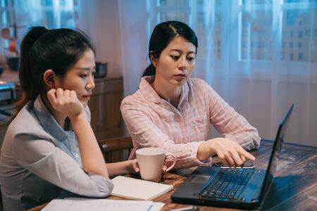young beautiful asian girl in nightwear helping woman worker best friend to complete her project on laptop computer. female point to screen teaching roommate in midnight trying to solve problem.の写真素材