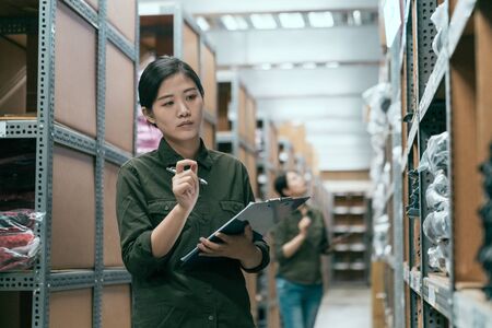 woman worker doing stocktaking of products in cardboard box on shelves in warehouse using clipboard and pen. female staff checking stock in factory stockroom. girl working in storehouse with coworkerの写真素材