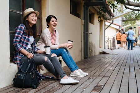 two happy young girls friends relax sitting outdoor leaning on old house window in little village. female japanese travelers visit in taiwan taipei small town with beautiful sunset light real momentの写真素材