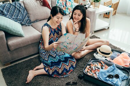 high angle top view of two happy young girl friends sitting on floor on carpet by messy suitcase. group of cheerful sisters plan search talk discuss tourism attraction on paper map packing luggageの写真素材
