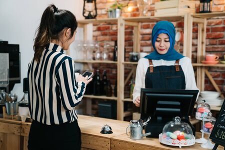muslim woman worker takes customer order in coffee shop. young business office lady morning routine in cafe bar buy breakfast using smart phone pay counter. islam girl waitress in headscarf smilingの写真素材