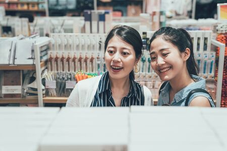 two happy asian women with curious face choosing markers for drawing in art shop. young girls buying shopping in stationery store for pen. high angle view with smiling female best friends indoors.の写真素材