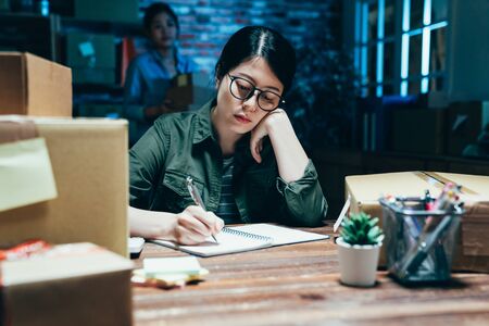 tired Asian business lady in glasses look at customer address on pastal package writing down on...