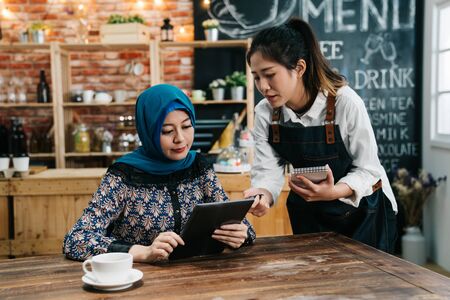 Modern islam businessman reading menu on tablet in cafe bar while waitress recommending guest something special. young girl waitress taking order point to touch pad in coffee shop giving advice.の写真素材