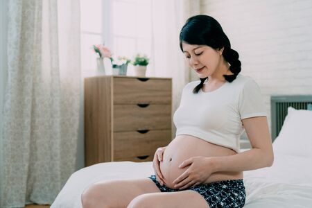 Happy Asian pregnant woman in braid touching her bare belly with care in bedroom.