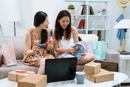 two asian chinese young women unpacking parcels at home sitting on couch surrounding by boxes and shopping bags. girls friends with credit card and laptop computer buying purchase on internet saleの写真素材