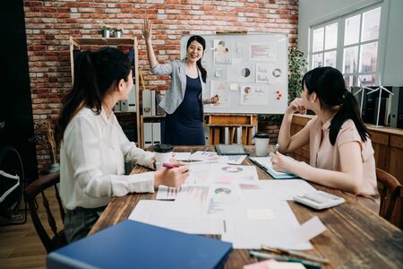 group of creative female people doing brainstorming meeting in modern studio. three women colleagues in board room discussing project. pregnant manager lady standing at white board give presentationの写真素材