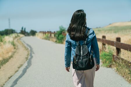 back view of asian woman traveler happy relaxing in grassland outdoor under blue sky with sunlight. young girl backpacker walking on road path way between mountain forest tree and wood vintage fenceの写真素材