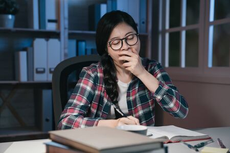 Young woman student at home desk reading and yawning tired at night with pile of books. exhausted college girl preparing exam in midnight stay up late hard working. university education conceptの写真素材