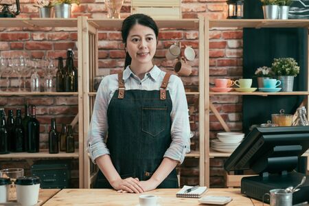Young female barista standing behind bar counter in cafe smiling and looking at customer while their order is ready on table. woman waitress in apron waiting for client to take cup of coffee.の写真素材