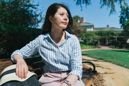 Girl sunbathing on park bench. patient asian female traveler waiting for bus in front of house and sitting on wooden chair. elegant lady relax with backpack and sun hat in park surrounding by grass.の写真素材