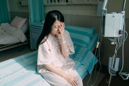 asian korean woman patient in hospital room sitting on bed sick and injured feeling sad and depressed. illness lady with saline drip bag on hand covering face. sunshine through window bright indoorの写真素材