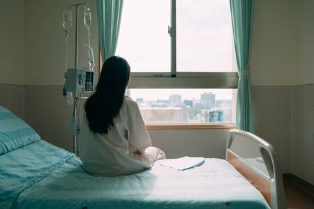 back view of lonely young asian woman sitting on bed in hospital ward. depressed female patient stay in room with body check result paper beside. sad lady in pajamas looking out window indoorの写真素材
