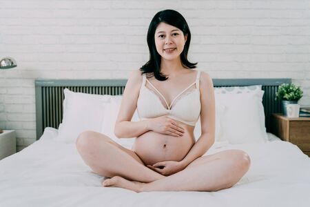 future mommy in sexy lingerie sitting on white bed. cozy bedroom interior for pregnant woman. attractive anticipation mom relax at home in comfort bra and panties face camera smiling hold  bellyの写真素材