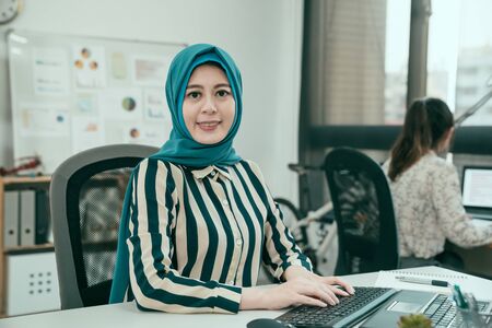 vintage style photo of Arabian woman with happy smile face camera while working in office. charming islam lady worker in blue hijab typing on computer in studio. female collegaue sit in backgroundの写真素材