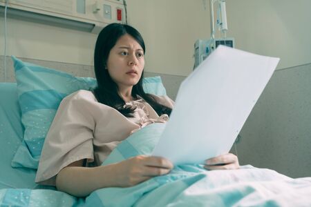 worried asian woman patient shocked to read results of medical examination. illness young girl sitting on bed in hospital room holding document of body situation. frowning lady look at paper in wardの写真素材