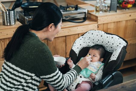 happy korean mother holding bow and spoon is feeding baby at lunch time. loving asian female parent is saying "ah"" while giving her kid solid food at home.  の写真素材