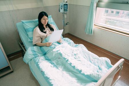 unhappy woman patient reading paper of bad test result while lying in hospital bed in room. cancer treatment oncology concept. depressed illness lady relax in recovery ward in sunshine clinic.の写真素材