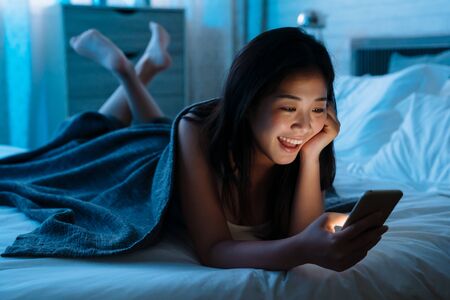 Happy asian japanese teen lying in front in bed at home with smartphone. Young joyful woman watching funny video on mobile phone in bedroom at night and laughing. relax girl shopping online indoorsの写真素材