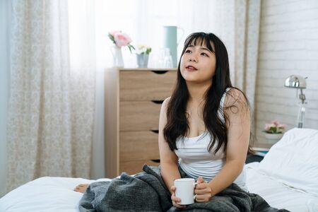 Beautiful asian korean girl at cozy bright bedroom drinking cup of tea in morning. charming woman sitting in comfort bed holding mug with hot cocoa. cute female relax in home early time indoors.の写真素材