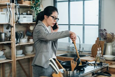 busy korean female worker holding spoon is frying egg and dealing with work on phone in the morning. asian businesswoman using spatula and pan is calling her boss and cooking breakfast for family.の写真素材