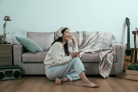 young asian woman listens to songs from mobile phone with earphones sitting at home wooden floor in living room isolated on white background. smiling relax lady in sweater enjoy music by headphonesの写真素材