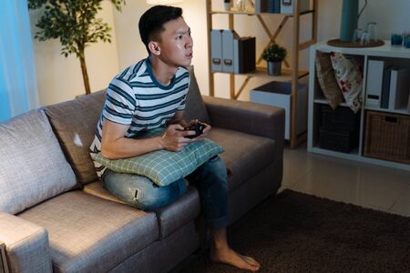 Young asian chinese man addicted to video gaming at dark home. concentrated handsome guy in casual wear stay up late house living room. male sitting on sofa holding joystick remote in evening.の写真素材