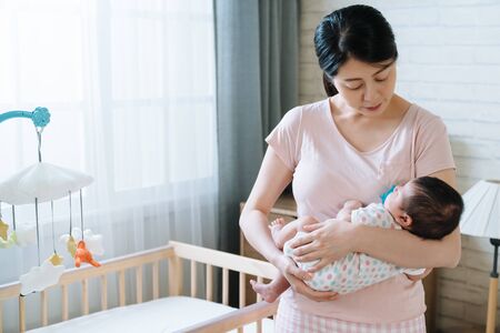 korean woman holding her baby girl is walking her baby to sleep in bedroom. asian new mom watching...