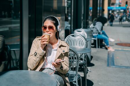 Pretty woman relax outdoors street coffee shop sit at table listen music in headphones and using mobile phone. happy carefree female in sunglasses drinking paper cup beverage under sunshine in cafeの写真素材