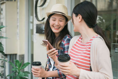 Sunny summer day travel lifestyle. women friends standing outdoors with smartphone and disposable paper cup in hands. two girls laughing and sharing cellphone together while drinking coffee by windowの写真素材