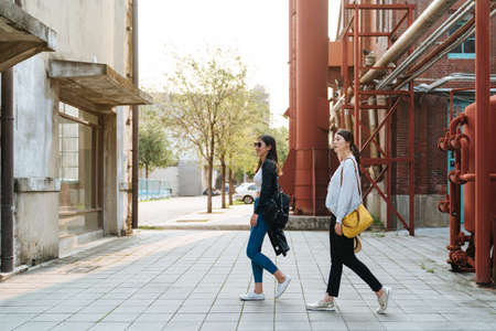 side view full length of two asian japanese women friends relax sightseeing in old factory tourist attraction. group of sisters in fashion wears walking in street while visiting vintage red building.の写真素材