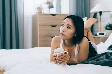 blurred background pretty asian girl lying prone on bed with coffee is feeling bored. portrait of korean young woman at a loose end in her bedchamber.の写真素材