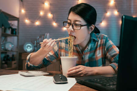 elegant businesswoman eating instant noodles at dining table while over work on project with laptop computer and calculator.の写真素材