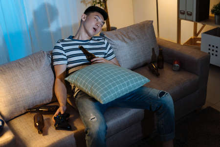 handsome lazy asian korean man sleeps on comfort couch in evening living room after drinking bottles of beer and playing video game. tired guy fell asleep in sofa at late night indoors in apartmentの写真素材