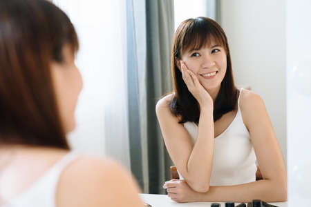 beautiful japanese girl is looking at her reflection in front of mirror with pleasant expression....